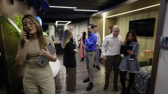 A confident business woman talks on her phone and holds a tablet in a modern office. Her colleagues engage in discussions and networking in the contemporary workspace.