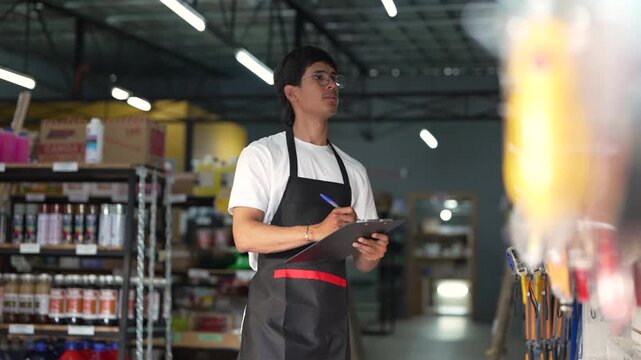 Young male worker in an apron managing stock and writing on a checklist in a large retail warehouse