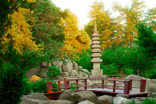 Serene Japanese garden with a wooden bridge and stone pagoda surrounded by vibrant green and yellow trees and large rocks in Ukraine Kyiv park named Kioto