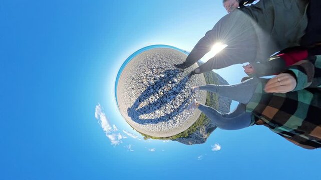A rotating tiny planet effect showing a couple walking along its surface, with a pebbled beach, turquoise water, green cliffs, and a vivid blue sky surrounding them