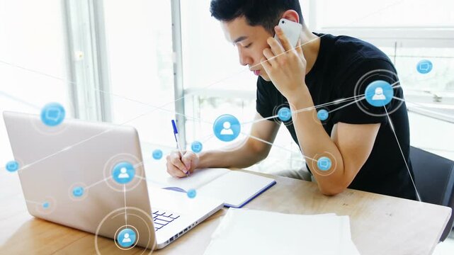 Animation of profile icons over asian man taking notes while talking on smartphone at office