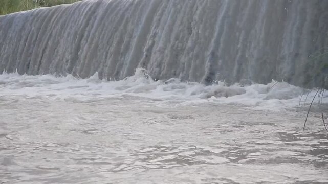 Muddy river water flowing over a small dam, creating a textured cascade with splashing foam and dynamic motion, ideal for flood, environment, and water management concepts.