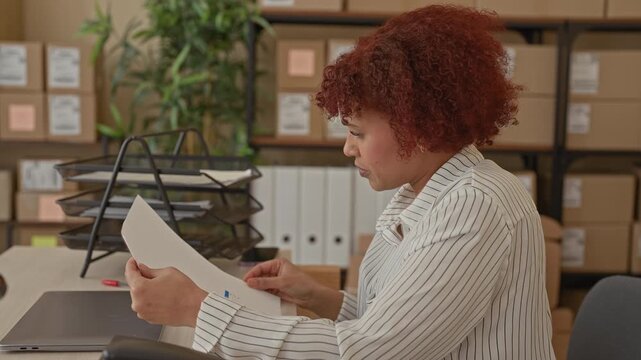 Woman holding documents raises arms in a building filled with cardboard boxes and parcels, pointing to papers; joy success.