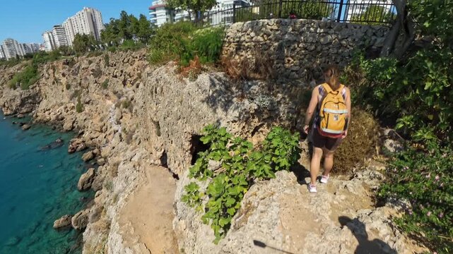 A Woman walks along a narrow cliffside path above the ocean, surrounded by greenery. The video is taken with a wide-angle lens, emphasizing the steep drop and coastal view.