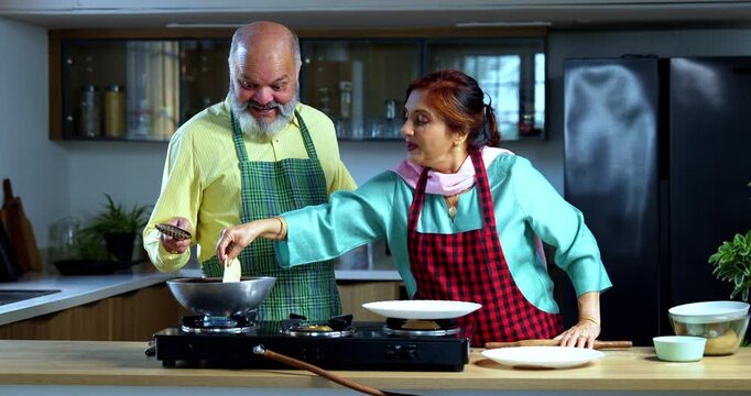 Retired senior Indian Asian old age couple together frying puri in a modern kitchen; showcasing teamwork, traditional food preparation, and happy lifestyle in a contemporary home setting.