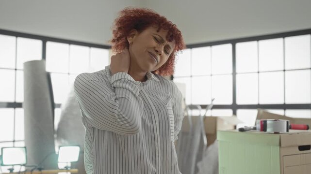 Woman with hand on neck and closed eyes among moving boxes and packing tape in building; moving day discomfort.