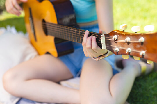 Strumming Asian woman playing acoustic guitar cross-legged on picnic blanket in park, with sunlight