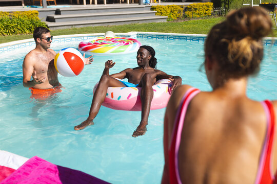 Diverse friends playing in backyard pool, tossing beach ball beside donut float with colorful rings