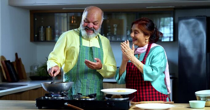 Retired senior Indian Asian old age couple together frying puri in a modern kitchen; showcasing teamwork, traditional food preparation, and happy lifestyle in a contemporary home setting.