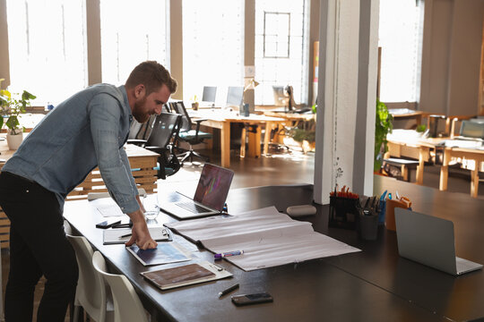 Male architect studying blueprint on table with laptop tablet smartphone in office, copy space