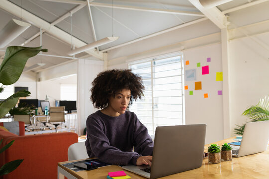 African American woman typing on laptop at open-plan office desk with phone and sticky notes