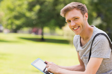 Man smiling while tapping tablet on park grass wearing grey t-shirt and backpack strap, copy space © wavebreak3