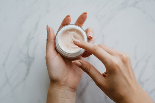 Hands holding and scooping cream from jar on marble surface