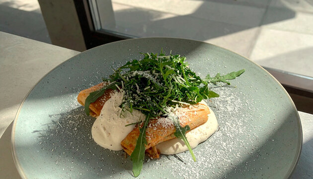 Savory crepes topped with arugula, white sauce, and grated cheese on a grey plate