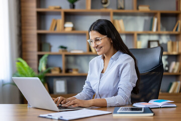 Young indian woman wearing glasses cheerfully working on a laptop at her desk in a contemporary...