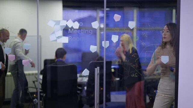 Business professionals actively brainstorm and collaborate on a glass wall using sticky notes and markers in a modern office.