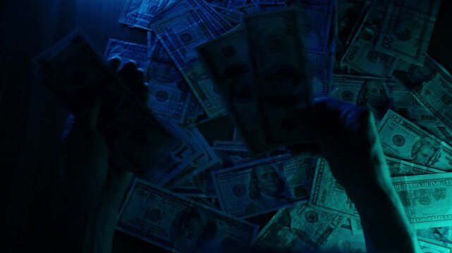 A woman's hands pick up US dollar banknotes from a scattered pile on a table under dramatic blue lighting, evoking a sense of urgency and action