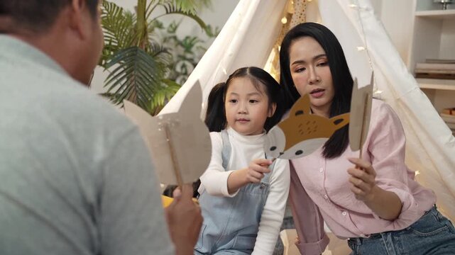 Happy Asian family plays together in an indoor teepee tent with fairy lights and a Christmas tree. Mother and young daughter hold up a cute fox paper mask and a plain paper mask for the father