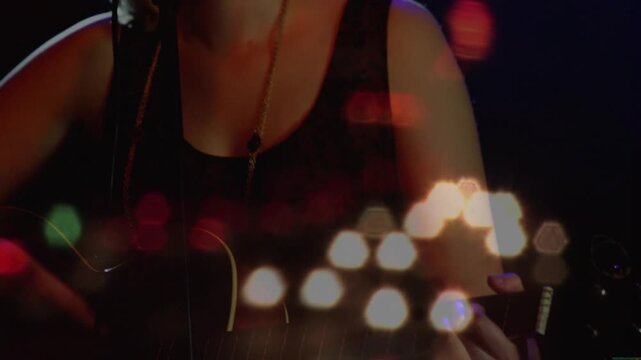 Camera pulling back female singer fretting strumming guitar leaning into mic singing amid bokeh