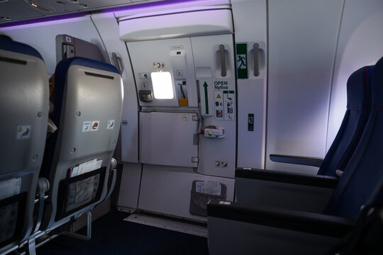 Interior of a commercial aircraft showing the emergency exit door, cabin attendant seat, and passenger rows with blue upholstery.