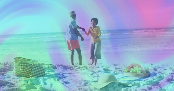Twirling couple on sandy beach with waves, woman in yellow, man in blue shirt, picnic basket