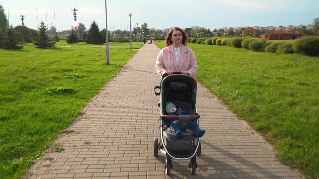 a mother in a pink coat walks with her newborn baby sleeping in a stroller down a long brick path in a green park, with city buildings visible in the autumn background.