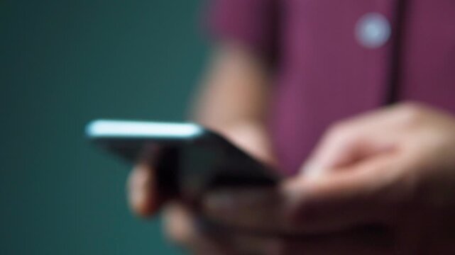 Abstract out of focus footage of a person using a mobile phone. Soft bokeh lighting highlighting hands typing and scrolling on a smartphone screen.