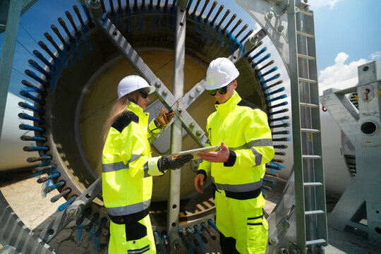 Engineers in high-visibility jackets review plans on a tablet near wind turbine components