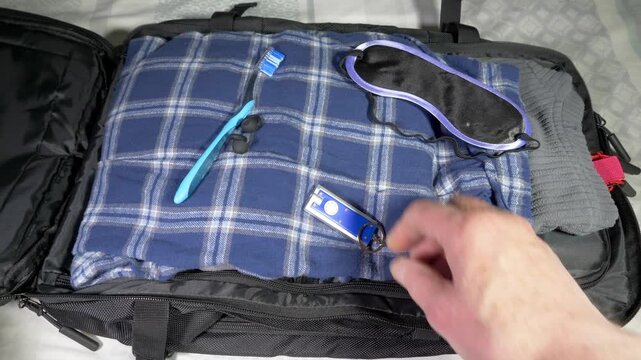 Closeup overhead shot of a man&rsquo;s hands unzipping a travel backpack on a bed cover, and removing a small torch from sleep clothing and other related items.