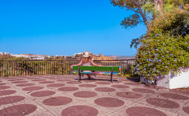 A woman wearing a sun hat sits on a colorful bench at the Mirador de Abades viewpoint with panoramic views of the Castle of the Dukes and Arcos de la Frontera, Cádiz, Spain.