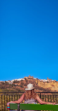 A woman wearing a straw hat sits on a bench at the Mirador de Abades, with a panoramic view of the Castle of the Dukes and the white town of Arcos de la Frontera, Cadiz, under an immense blue sky.