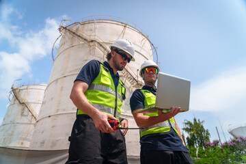 Industrial workers in safety vests and hard hats review data on a laptop near storage tanks © FotoArtist