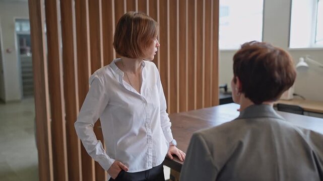 Two women standing and talking in front of a wooden divider in a modern office lounge. Discussion, standing, business, office, teamwork.