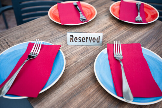 Close-up of a reserved restaurant table set for four with blue and red plates, red napkins, and forks on a wooden table.