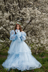 Fototapeta premium young woman in blue dress standing under blooming tree with floral headpiece, soft spring light, delicate white blossoms, dreamy romantic garden portrait