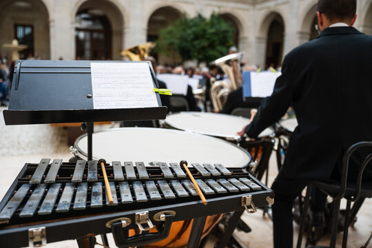 Rear view of a percussionist in an orchestra, standing with mallets at a xylophone and timpani in an arched courtyard.