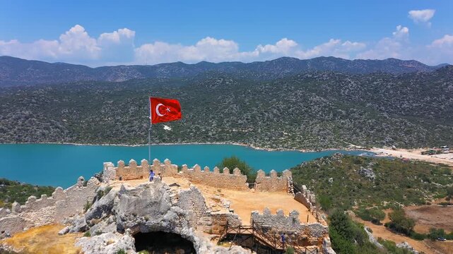 Turkish flag waving over Simena Castle battlements with turquoise Kekova lagoon and forested mountains stretching behind, Antalya, Turkey