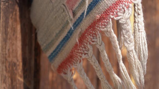 Macro shot of the frayed edge of a handwoven ethnic Turkish kilim rug, showcasing detailed fabric texture, colorful stripes, and braided tassels hanging against a rustic wooden background in sunlight