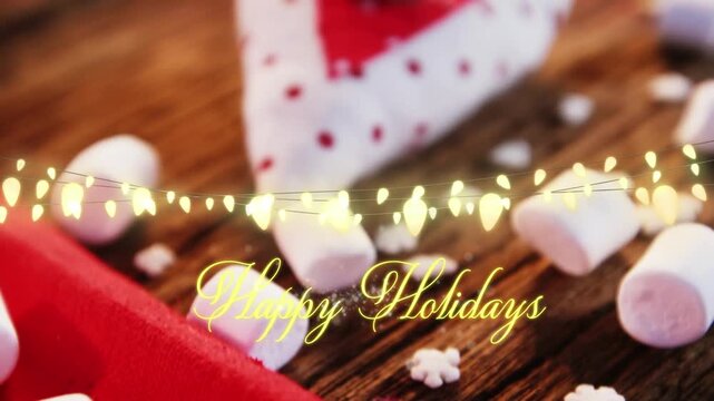 Animation of text, happy holidays, in yellow, over string lights, marshmallows and decorations