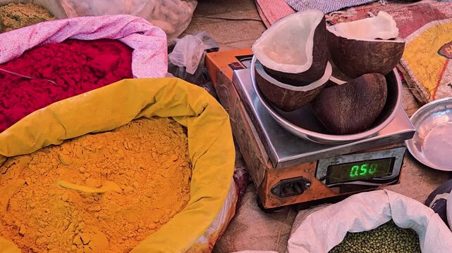 A vibrant market stall displays halved dry copra coconuts being weighed, showing 0.504kg on a digital scale. They're surrounded by open sacks of bright turmeric & red powders in authentic marketplace.