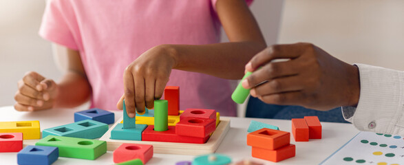 Fototapeta premium Two children engage in play with various colorful shapes on a table. One child uses blocks while the other adds pieces to the structure. The room is well-lit and inviting.