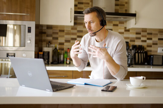 Man in Headset Having Online Meeting in Modern Kitchen. Focused man wearing a headset with microphone while talking during a video conference on his laptop. Home office and remote work
