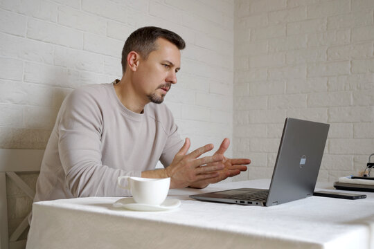 Serious Man Gesturing During Video Conference at Home Table. Focused man sitting at table, explaining something during an online meeting on his laptop. Freelance, work from home