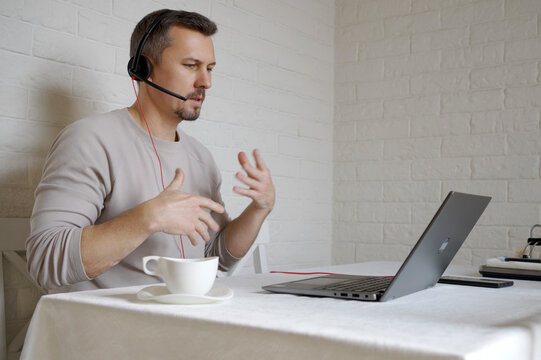 A Man in Headset Leading Online Meeting from Home Office. A focused man wearing a headset gestures while talking during a video call on a laptop. Remote work, and freelance concept