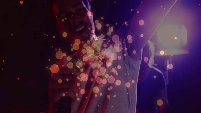 Music performer singing into mic, purple lights shifting causing chest sparkles by bandmate on keys