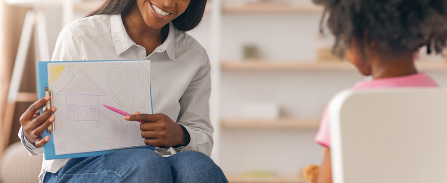 A teacher is smiling as she shows a drawing to a young student sitting in front of her. The scene takes place in a classroom with shelves in the background.