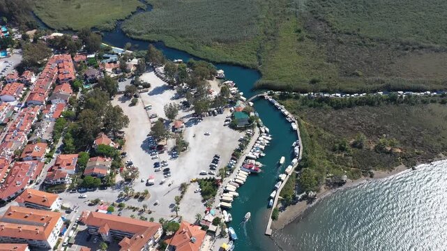 Aerial view of Azmak River marina with boats moored along natural channel, reed wetlands and red-roofed Akyaka town beside the sea, Mugla, Turkey