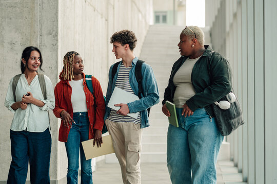 Diverse students walking, talking, and interacting in a modern university hallway, carrying books and laptops on campus