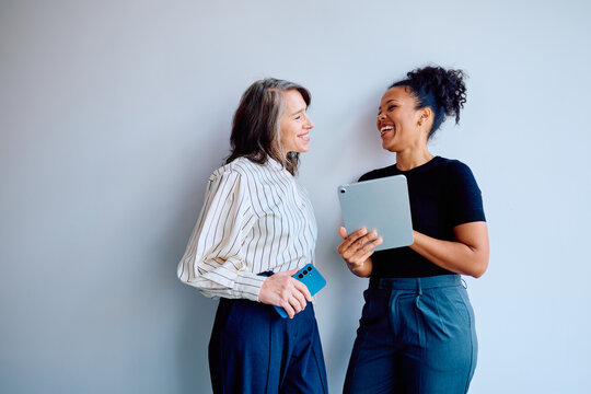 Two diverse business women laughing, communicating, and interacting while using a digital tablet and smartphone in an office setting