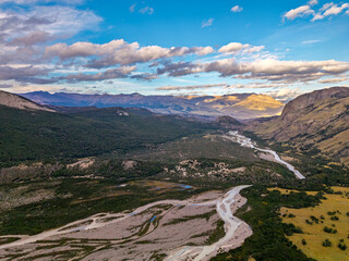 Aerial panoramic view of a wide braided river valley winding through the mountainous landscape of Patagonia, South America, under a dramatic cloudy sky.